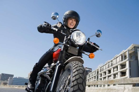 Une femme souriante en veste et casque noirs conduit une moto roadster rouge et noire en milieu urbain sous un ciel bleu, profitant pleinement des avantages de la moto au quotidien.