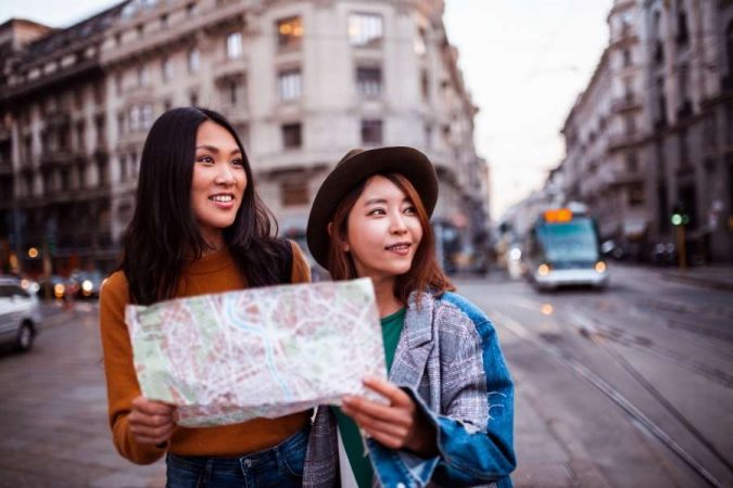 Deux jeunes femmes souriantes avec une carte en main dans une rue animée d'une grande ville européenne, partant à la découverte de la culture d'un pays.
