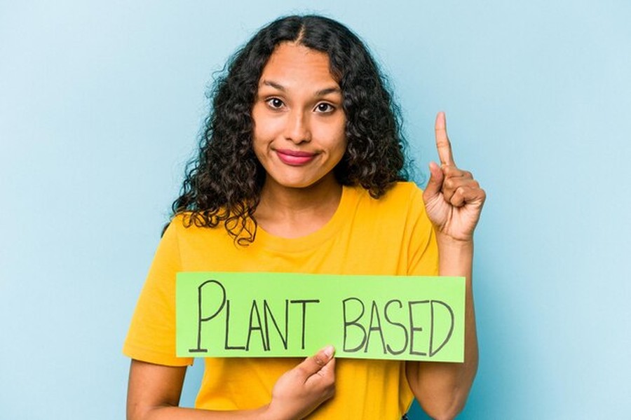 Une jeune femme en t-shirt jaune tenant une pancarte verte 'PLANT BASED' tout en expliquant comment appliquer l'affichage environnemental obligatoire sur vos fiches produits en 2026.