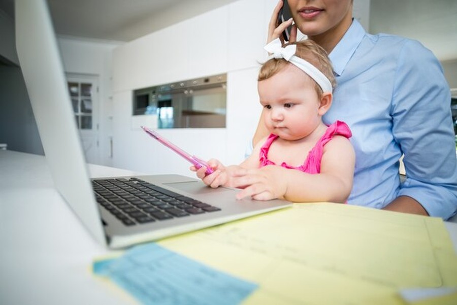 Une mère travaille sur un ordinateur avec son bébé, soulignant l'importance de savoir comment protéger l'identité numérique de son enfant dès la naissance.
