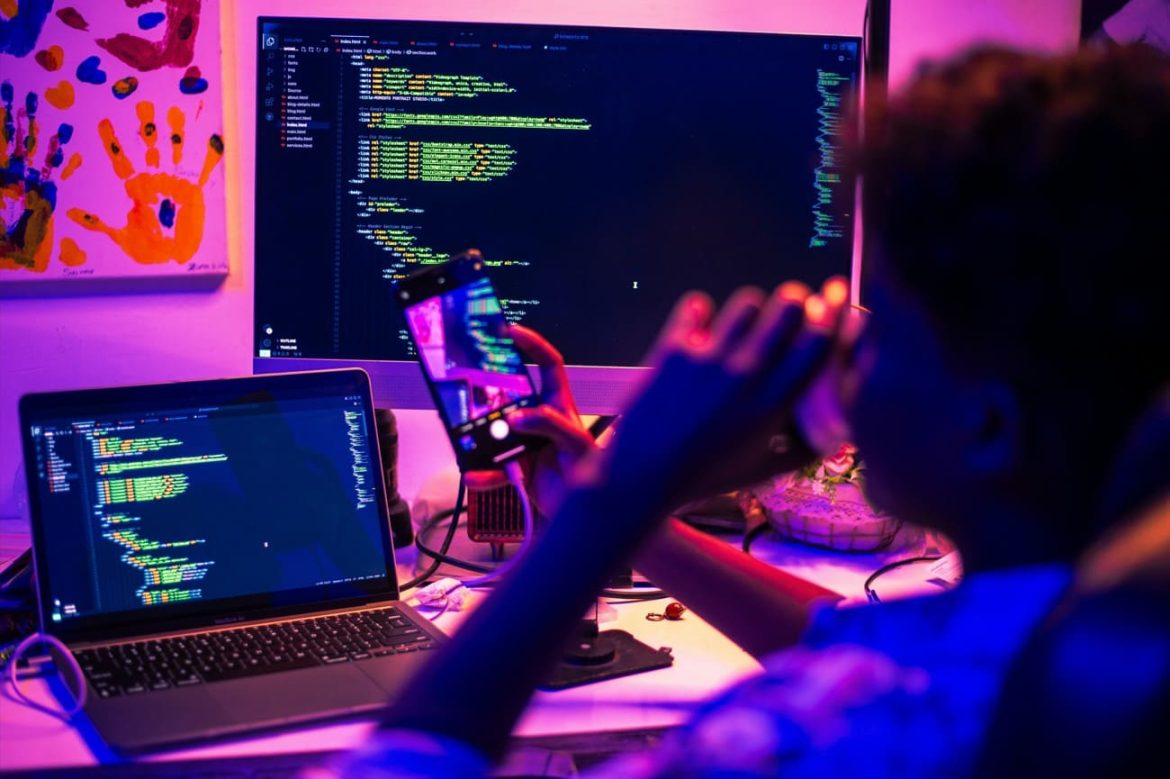 Plan d'ensemble d'un élève de dos travaillant sur un ordinateur portable avec un grand écran de programmation, illustrant l'environnement propice à l'l'IA et la réussite des élèves.