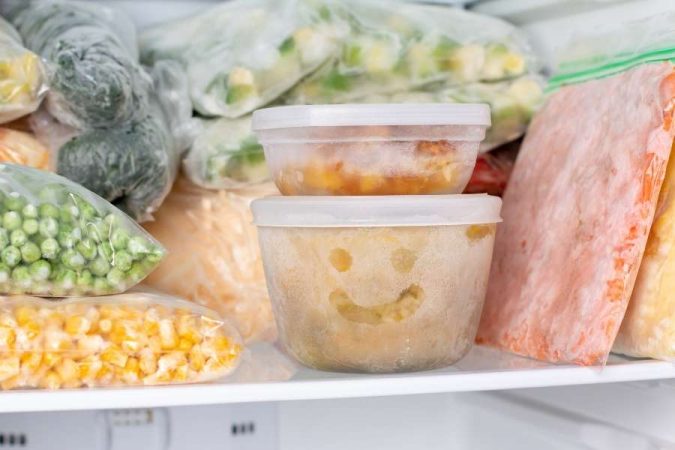 Des boîtes de conservation empilées et des sachets de légumes congelés dans un congélateur.
