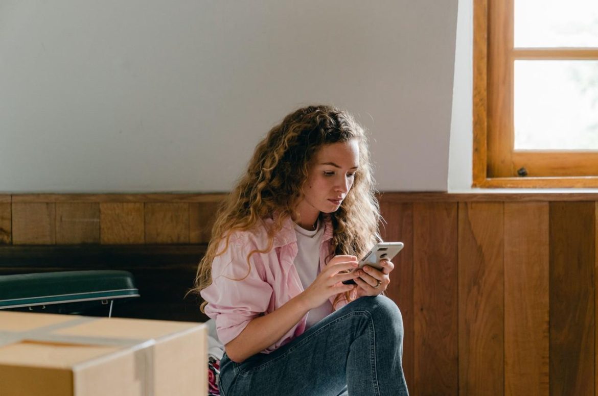 Une jeune femme assise au milieu de cartons utilise son smartphone pour consulter une application déménagement.