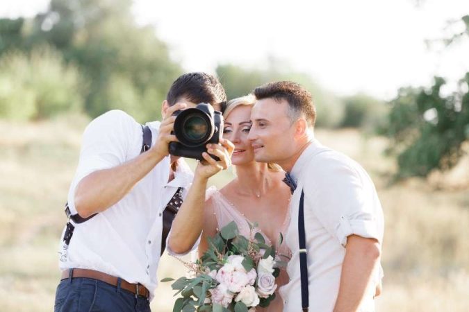 Un photographe de mariage professionnel montre une photo sur l'écran de son appareil aux mariés souriants en extérieur.