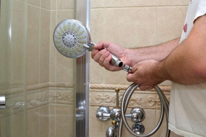 Un homme tient un pommeau de douche chromé dans une main et l'extrémité dévissée d'un vieux flexible de douche métallique dans l'autre.
